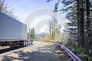Big rig classic American bonnet semi truck transporting cargo in refrigerator semi trailer turning on the mountain road with trees