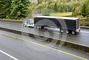 Big rig classic day cab semi truck transporting cargo in covered black semi trailer running on the wet slippery road