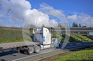 Big rig classic high cab semi truck with storage boxes on the back side transporting empty flat bed semi trailer running on the