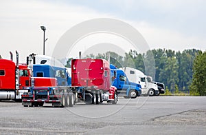 Big rig classic red semi truck with empty flat bed semi trailer driving on the industrial parking lot with another semi trucks