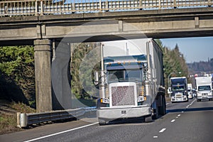 Big rig classic semi truck with covered semi trailer driving on the road under the bridge in front of semi trucks convoy