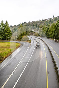 Big rig classic semi truck with covered semi trailer running on the wet divided turned road with green trees on the hill