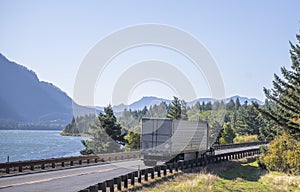 Big rig classic semi truck transporting goods in refrigerator semi trailer running on the road along river in Columbia Gorge area