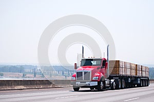 Big rig day cab red semi truck with flat bed trailer transporting lumber wood by wide multiline highway