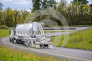 Big rig day cab semi truck with bulk semi trailer turning on the highway entrance with green trees on the sides