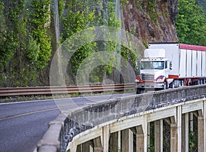 Big rig day cab semi truck transporting bulk semi trailer driving on mountain road with rock wall and bridge