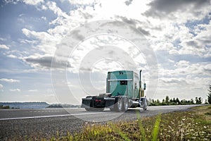 Green classic big rig semi truck tractor driving on the road with cloudy sky to warehouse for pick up the loaded trailer