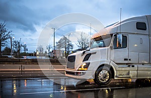 Big rig long haul white semi truck transporting goods running on the wet road with rain at night time