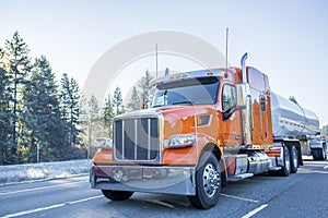 Big rig orange classic American semi truck transporting liquid cargo in tank semi trailer moving on the winter frosty road