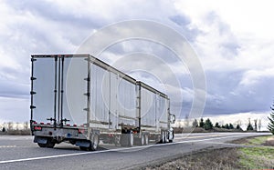 Big rig powerful semi truck transporting goods in two long semi trailers running on the straight road