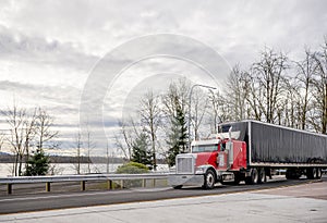 Big rig red classic bonnet semi truck transporting cargo in covered rubberized black semi trailer driving on the road intersection