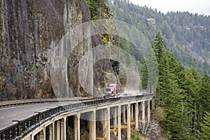 Big rig red semi truck with flat bed semi trailer driving on the wet narrow highway road with bridge builded around mountain rock