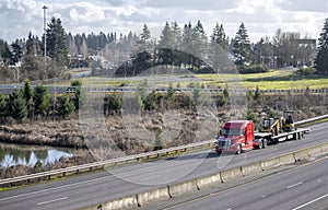 Big rig red semi truck with low roof transporting small tractors on step down semi trailer running on the divided highway road