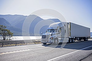 Big rig semi truck with chrome accessories transporting frozen cargo in refrigerated semi trailer moving on the road along river