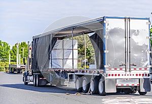 Big rig semi truck with open covered semi trailer unloading commercial cargo on warehouse parking lot