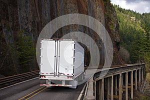 Big rig semi truck with refrigerator semi trailer moving on bridge on side of big rock wall in Columbia Gorge