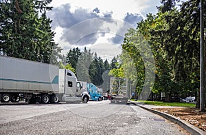 Big rig semi truck with shiny semi trailer driving out of rest area parking lot with different resting semi trucks standing in row