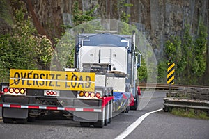 Big rig semi truck with sign oversize load behind flat bed semi trailer driving on the mountain road with rock wall on the side