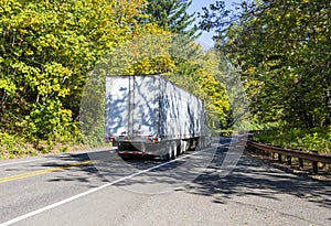 Big rig semi truck tractor transporting cargo in dry van semi trailer running on the winding road in forest