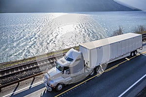 Big rig semi truck transporting cargo in dry van semi trailer running on sunny road along wide river