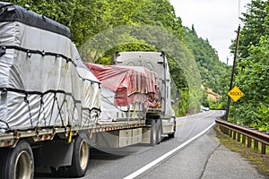 Big rig semi truck transporting covered and tightened commercial cargo on flat bed semi trailer running on the winding road at