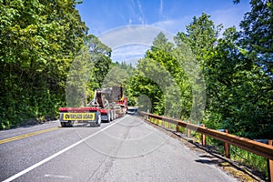 Big rig semi truck transporting oversize load on the step down semi trailer movig on the road with green trees