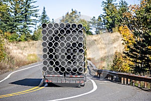 Big rig semi truck transporting plastic pipes on the flat bed semi trailer running on the winding road with hills and trees