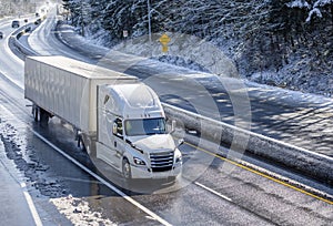 Big rig white bonnet semi truck with dry van semi trailer moving on the winding winter road with wet surface and snow