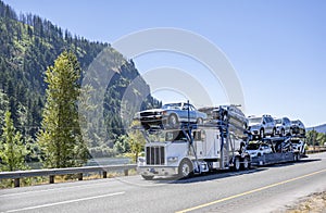 Big rig white classic car hauler semi truck transporting cars on the modular two level semi trailer driving on the one way road