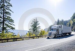 Big rig white classic semi truck transporting cargo in refrigerated semi trailer on winding autumn road