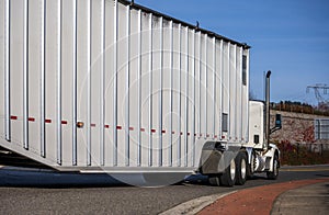 Big rig white day cab semi truck transporting long covered bulk semi trailer driving on the local road