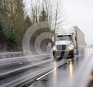 Big rig white diesel semi truck transporting container with commercial cargo running on the wet road at raining weather