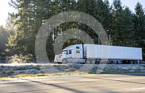 Big rig white long haul semi truck with dry van semi trailer standing on winter parking lot with evergreen trees on background