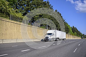 Big rig white semi truck with dry van semi trailer running on the wide multiline highway road with concrete wall and trees on the