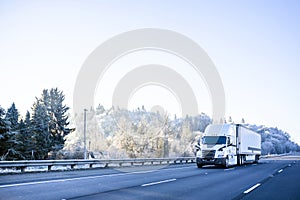 Big rig white semi truck with refrigerator semi trailer driving on the winter highway with snow and frozen trees on the hillside