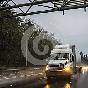 Big rig white semi truck with semi trailer driving on wet raining evening road with turned on headlight and reflection