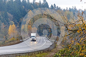 Big rig white semi truck with trailer and turned on headlight driving on the winding wet road in raining autumn weather with