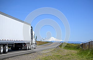 Big rig white semi truck transporting cargo in dry van semi trailer running on the narrow road with snow mountain ahead