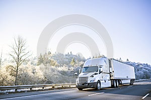 Big rig white technological semi truck with reefer semi trailer going on the winter road with frost trees on the hill photo