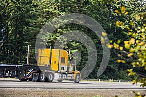 Big rig yellow semi truck with step down semi trailer running on the highway with green and autumn trees