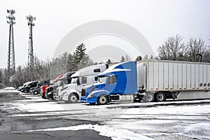 Big rigs semi trucks with different semi trailers standing for truck driver rest on the winter truck stop parking lot with snow