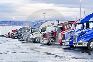 Big rigs semi trucks with semi trailers standing in row on truck stop parking lot with wet surface waiting for the continuation