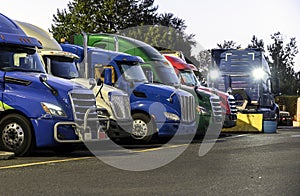 Big rigs semi trucks standing in row on truck stop parking lot on twilight with turn on headlight