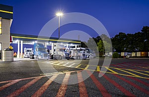 Big rigs semi trucks standing on the truck stop parking lot under a lighted awning at night