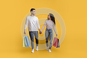 Big Sales. Happy Arab Couple Walking With Shopping Bags On Yellow Background