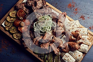 A big set of grilled meat with vegetables on big wooden plato or cutting board on gark wooden background. Tradicional