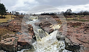 The Big Sioux River flows over rocks in Sioux Falls South Dakota with views of wildlife, ruins, park paths, train track bridge, tr