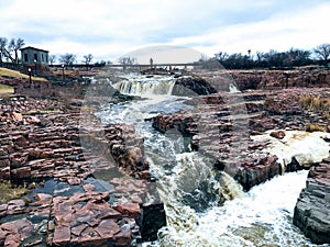 The Big Sioux River flows over rocks in Sioux Falls South Dakota with views of wildlife, ruins, park paths, train track bridge, tr