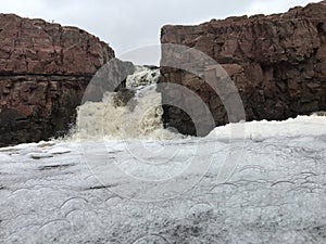 The Big Sioux River flows over rocks in Sioux Falls South Dakota with views of wildlife, ruins, park paths, train track bridge, tr