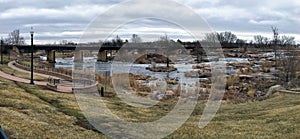 The Big Sioux River flows over rocks in Sioux Falls South Dakota with views of wildlife, ruins, park paths, train track bridge, tr